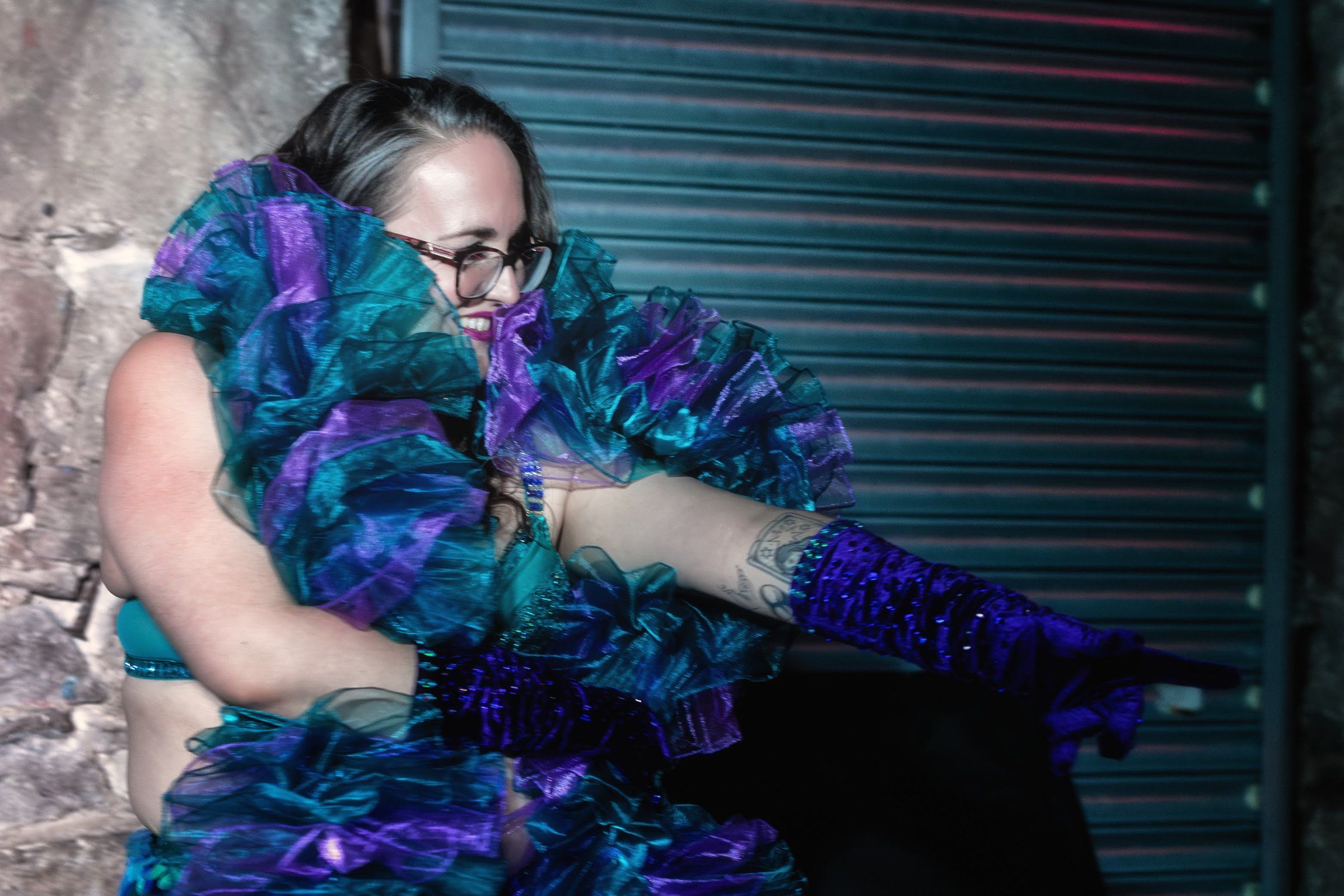Close-up of Esmeralda Fontaine in a teal and violet ruffled collar and long purple gloves, smiling and gesturing.