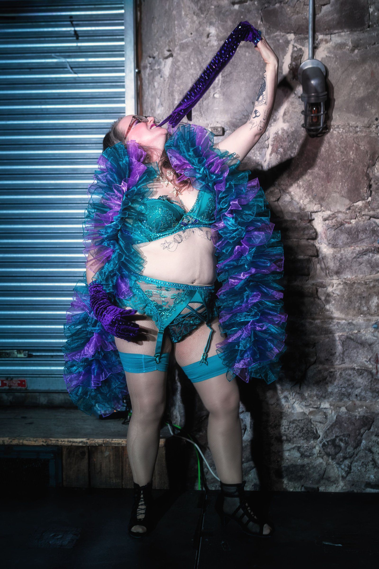 Esmeralda Fontaine poses in a teal and violet ruffled costume and purple gloves against an industrial roller door.