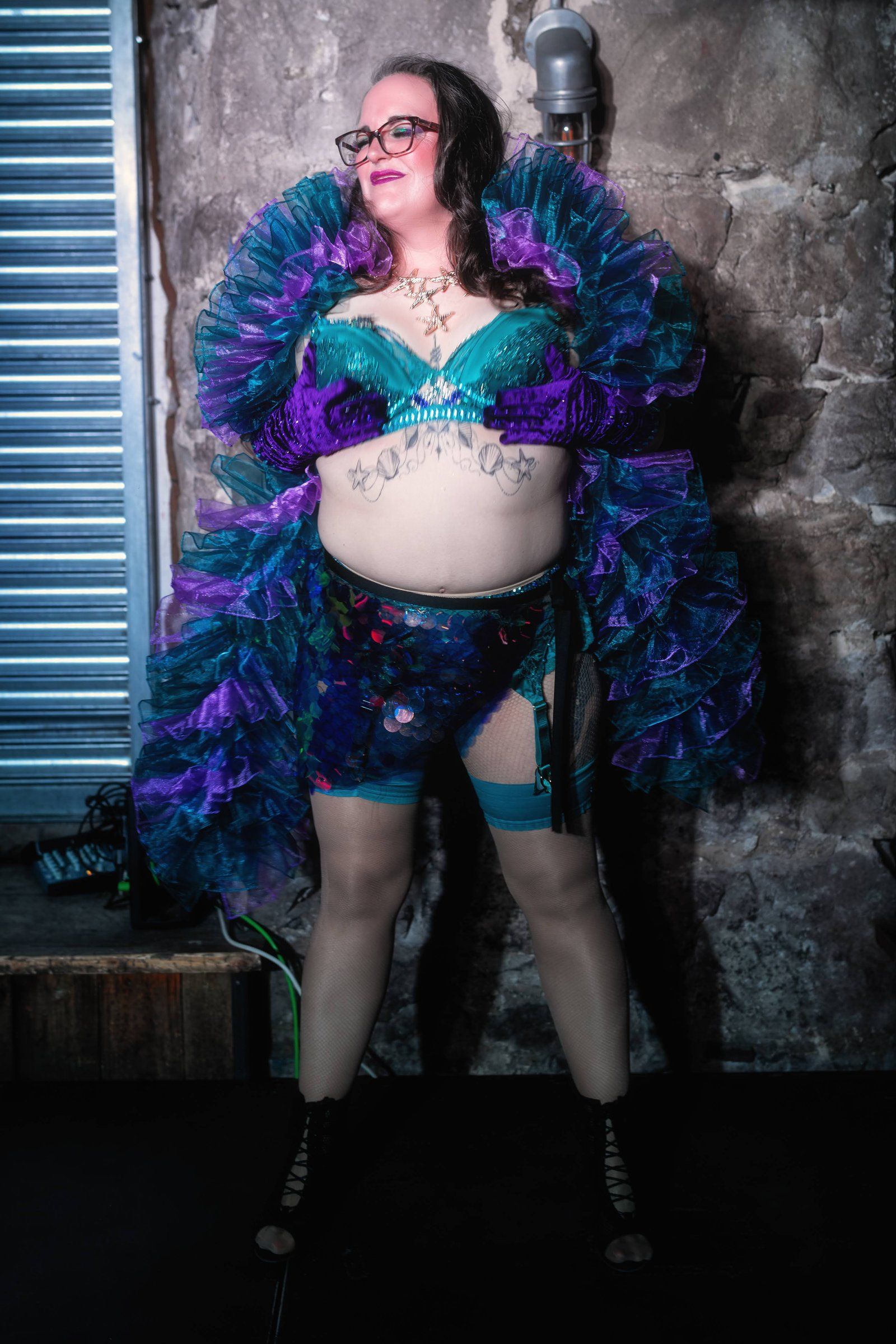 Esmeralda Fontaine stands against a distressed wall in a teal and violet ruffled costume, wearing glasses and looking upward.